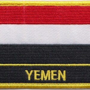 Può includere: Una toppa ricamata con la bandiera dello Yemen, con un design rosso, bianco e nero. La toppa ha un bordo giallo e la parola "YEMEN" in lettere gialle.