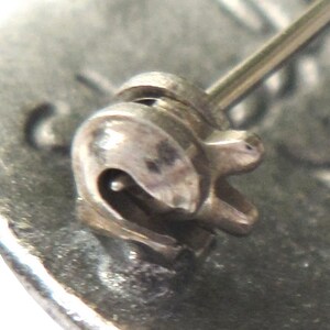 May include: Close-up of a silver pin back clasp with a spring-loaded mechanism. The clasp is designed to attach to a brooch or other jewelry piece.