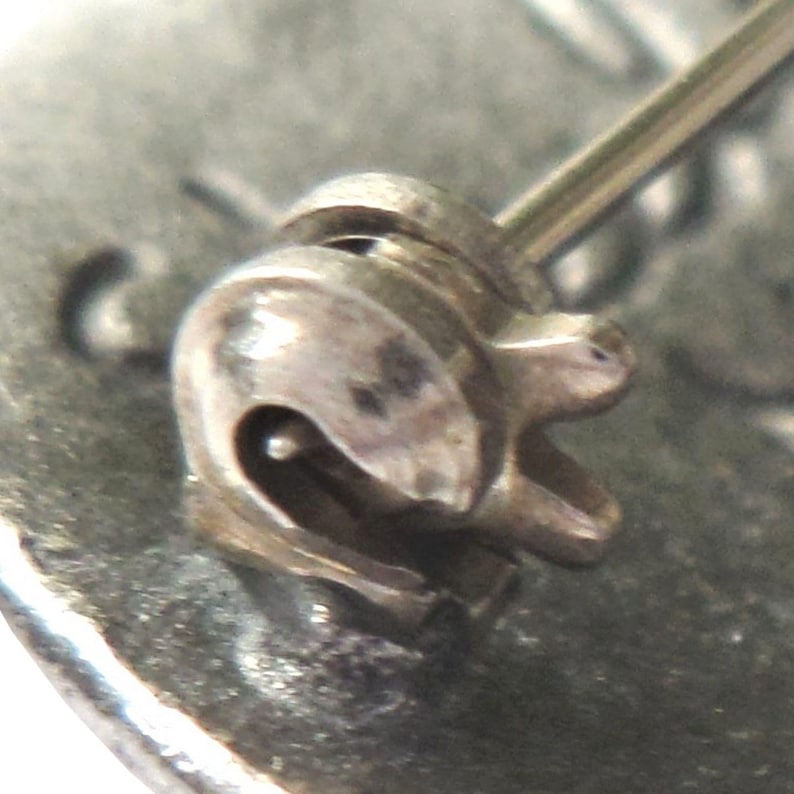 May include: Close-up of a silver pin back clasp with a spring-loaded mechanism. The clasp is designed to attach to a brooch or other jewelry piece.