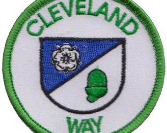 May include: A white circular patch with a green border. The text "CLEVELAND WAY" is written in green around the edge. Inside the circle is a white shield with a blue triangle and a white flower. A green acorn is on the right side of the shield.
