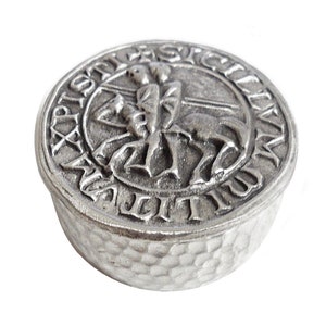 May include: A silver metal trinket box with a raised design of a knight on horseback. The box has a textured, hammered finish and the inscription "SIGILLVM  S.  IOANNIS  HIEROSOLYMITANI" around the edge.