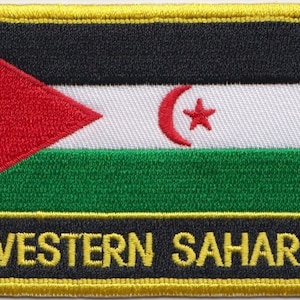 Peut inclure: Un écusson brodé jaune avec un drapeau noir, rouge, blanc et vert avec un croissant de lune rouge et une étoile. Le texte "WESTERN SAHARA" est écrit en jaune sous le drapeau.