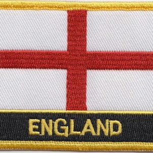 May include: A white embroidered patch with a red cross and the word "ENGLAND" in yellow lettering on a black background. The patch is surrounded by a yellow border.