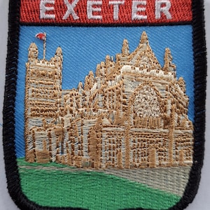 May include: Embroidered patch with a black border, a red banner with the word "EXETER" in white lettering, and a gold embroidered image of a building against a blue sky.