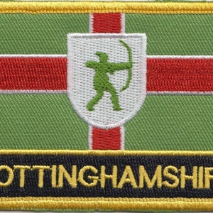 May include: A green, red, and white embroidered patch with a white shield containing a green archer figure. The patch has a black border and a gold border around the black border. The text "NOTTINGHAMSHIRE" is printed in yellow on a black background.