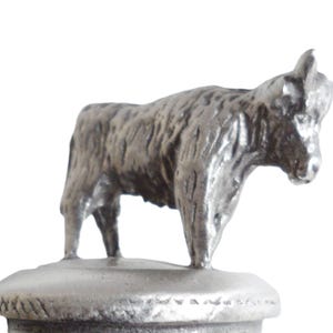 May include: A silver metal figurine of a Highland cow, standing on a round base. The cow is facing to the right and has a detailed, textured surface.