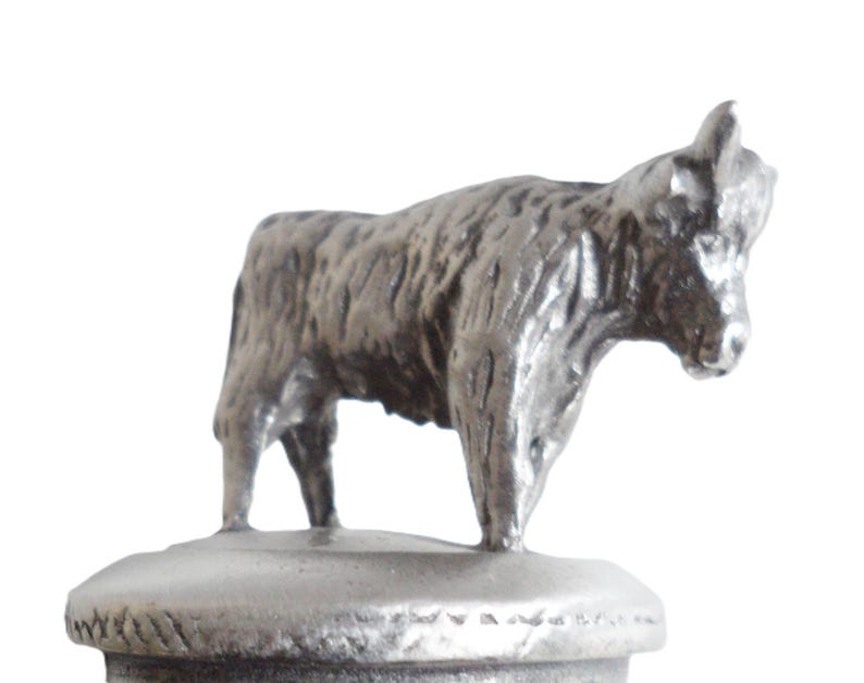 May include: A silver metal figurine of a Highland cow, standing on a round base. The cow is facing to the right and has a detailed, textured surface.