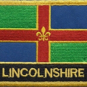 Op de afbeelding: Een geborduurde patch met een geel, rood, groen en blauw ontwerp, een gouden fleur-de-lis in het midden en de tekst "LINCOLNSHIRE" in gouden letters op een zwarte achtergrond.