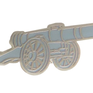 Op de afbeelding: Een zilverkleurige metalen pin in de vorm van een kanon met twee wielen. Het kanon is gedetailleerd met een zwarte omtrek en heeft een grijze achtergrond.