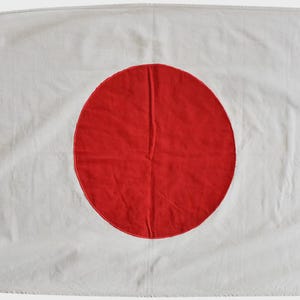 以下が含まれることがあります： 日本の国旗。白い長方形の旗の中央に大きな赤い円が配置されています。旗の左側には吊り下げるためのハトメが付いています。生地にはシワが見られます。