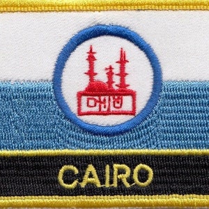 May include: A yellow, black, and blue embroidered patch with the word "CAIRO" in yellow lettering. The patch features a white background with a blue circle containing a red mosque.