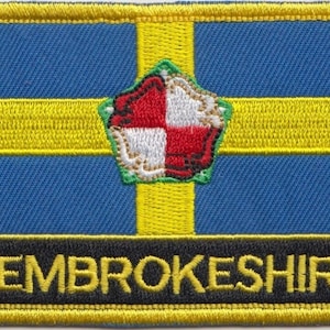 May include: A yellow embroidered patch with a blue background and a yellow cross. The cross has a red and white shield in the centre. The text "PEMBROKESHIRE" is embroidered in yellow on a black background at the bottom of the patch.