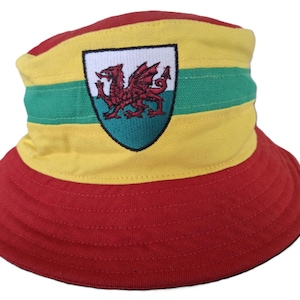 Puede incluir: Un sombrero de cubo rojo, amarillo y verde con un dragón galés bordado en la parte delantera. El dragón está en un escudo con fondo blanco.