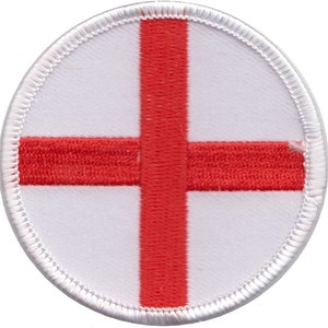 May include: A white circular patch with a red cross in the centre. The cross is made of red thread and is stitched onto the white fabric. The patch has a white border.