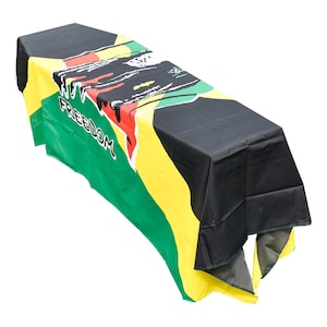 May include: A rectangular table cover in the colours of the Jamaican flag: green, yellow, and black. The word "FREEDOM" is printed in white on the green section. The cover is draped over a table, with the top and sides visible.