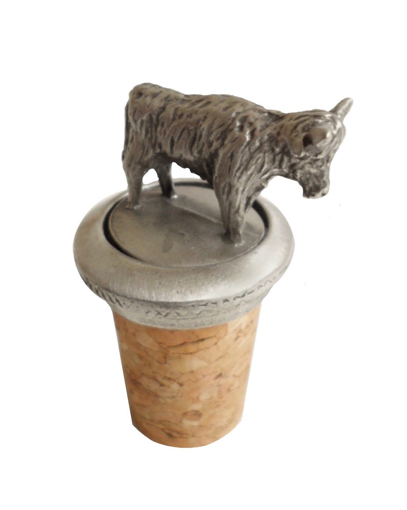 May include: A silver Highland cow bottle stopper with a cork base. The cow is standing on a round base with a textured rim.