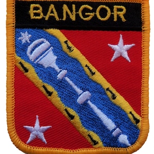 Peut inclure: Écusson brodé avec le mot "BANGOR" en jaune sur fond noir. L'écusson présente un champ rouge avec un motif diagonal bleu et jaune et deux étoiles blanches. Un emblème blanc traverse le motif bleu.