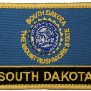 Könnte beinhalten: Ein gestickter Aufnäher in Blau und Gold mit dem Staatssiegel von South Dakota. Das Siegel zeigt eine Darstellung des Mount Rushmore und die Worte "The Mount Rushmore State". Der Aufnäher trägt auch die Worte "South Dakota" in goldenen Buchstaben auf schwarzem Hintergrund.