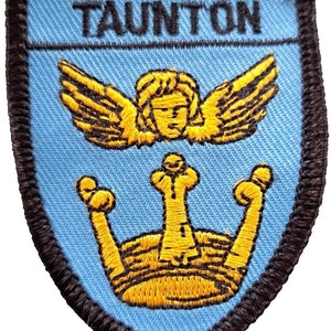 Peut inclure: Écusson brodé bleu clair avec le mot "TAUNTON" en haut. L'écusson présente un ange doré avec des ailes au-dessus d'une couronne dorée. L'écusson a une bordure noire.