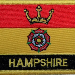 Może przedstawiać: Naszywka haftowana w kolorze żółtym, czerwonym i czarnym z koroną złotą, czerwoną różą z zielonymi liśćmi i napisem "HAMPSHIRE" złotymi literami.