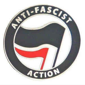 Puede incluir: Un pin circular blanco y negro con el texto "ANTI-FASCIST ACTION" alrededor del borde. El centro del pin presenta una bandera negra con una franja roja, simbolizando el movimiento antifascista.