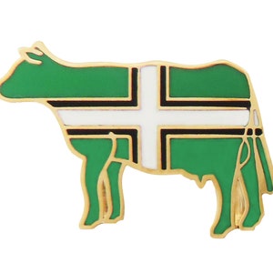 Könnte beinhalten: Eine goldfarbene Anstecknadel in Form einer Kuh mit einem grün-weiß gestreiften Design. Das Design ähnelt einer Flagge mit einem weißen Kreuz auf grünem Hintergrund.