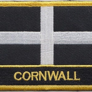Könnte beinhalten: Ein gestickter schwarzer und weißer Aufnäher mit einem weißen Kreuz auf schwarzem Hintergrund. Der Aufnäher hat einen goldenen Rand und das Wort "CORNWALL" ist unten in Gold gestickt.