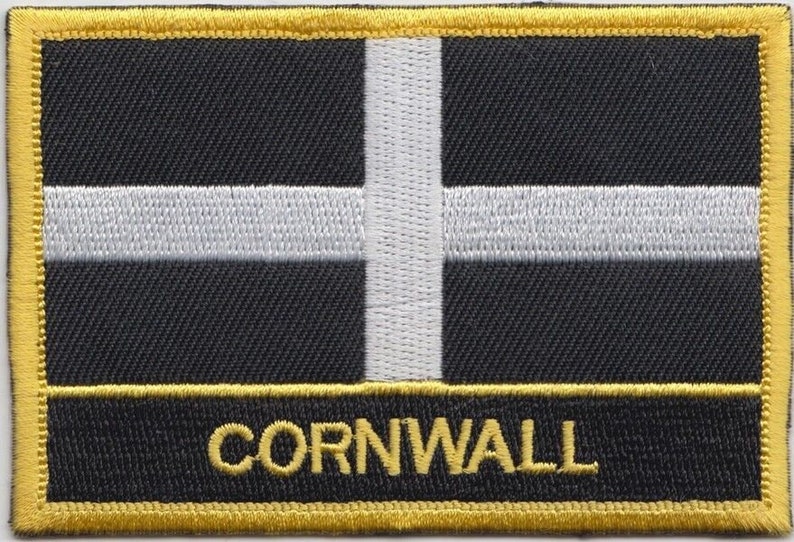 May include: A black and white embroidered patch with a white cross on a black background. The patch has a gold border and the word "CORNWALL" is embroidered in gold at the bottom.