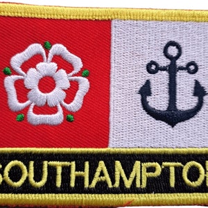 Puede incluir: Un parche bordado rojo y blanco con una rosa blanca y un ancla. El parche tiene un borde negro con la palabra "SOUTHAMPTON" en letras amarillas.
