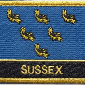 Pode incluir: Um patch retangular com o brasão de armas de Sussex. O design inclui seis martinetes dourados sobre um fundo azul, com uma faixa preta na parte inferior exibindo a palavra "SUSSEX" em letras douradas. O patch é bordado com costura dourada.