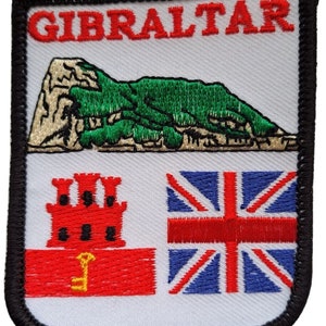 May include: A white embroidered patch with the word "GIBRALTAR" in red letters. The patch features a green and white mountain, a red flag with a white castle and a key, and a blue and red Union Jack flag.
