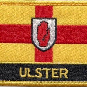 May include: Embroidered patch featuring the Ulster flag design. The patch has a yellow background with a red cross and a white shield with a red hand in the center. The word "ULSTER" is embroidered in gold on a black banner at the bottom.