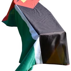 May include: A draped flag with a red top featuring a yellow starburst design and a black silhouette. The flag has green, white, and black sections. The flag is draped over an unseen object.