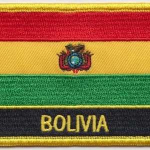 Pode incluir: Um remendo bordado com a bandeira da Bolívia. O remendo é vermelho, amarelo, verde e preto com a palavra "BOLIVIA" em letras amarelas. O brasão de armas nacional da Bolívia é bordado no centro.
