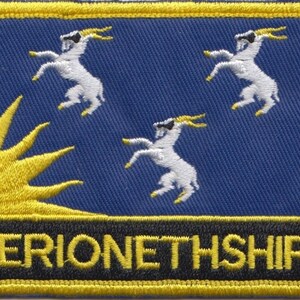Może przedstawiać: Naszywka haftowana w kolorze niebieskim i żółtym z trzema białymi kozami i żółtym słońcem. Tekst "MERIONETHSHIRE" jest napisany czarnymi literami na żółtym tle.