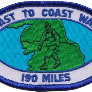 May include: White oval embroidered patch with blue trim. The patch features a green outline of a map with a hiker walking across it. The text "COAST TO COAST WALK 190 MILES" is embroidered in white.