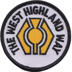 Puede incluir: Parche blanco bordado con costuras negras. El parche es redondo y presenta un cardo estilizado amarillo con el texto "The West Highland Way" en negro.