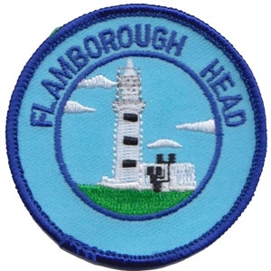 May include: Circular embroidered patch with a lighthouse design and the text "FLAMBOROUGH HEAD". The lighthouse is white with black details, set against a light blue backdrop with white clouds. The patch has a dark blue border.