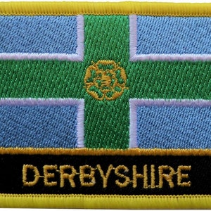 Può includere: Una toppa ricamata gialla con un disegno di bandiera blu, bianca e verde. La bandiera ha una rosa bianca al centro. Il testo "DERBYSHIRE" è ricamato in giallo su uno sfondo nero nella parte inferiore della toppa.