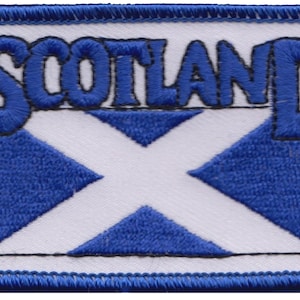 Puede incluir: Un parche blanco con una bandera escocesa bordada en azul y la palabra "SCOTLAND" en letras bordadas azules.