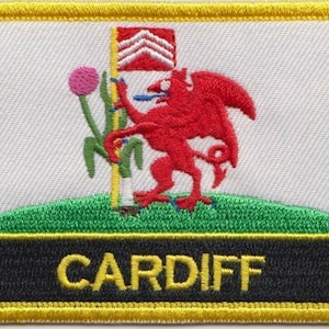 Könnte beinhalten: Ein weißer gestickter Aufnäher mit einem roten Drachen, einem grünen Hügel, einer rosa Blume und einer gelben Flagge mit roten Streifen. Der Text "CARDIFF" ist in Gelb auf schwarzem Hintergrund gestickt.
