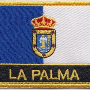 Pode incluir: Um remendo bordado azul e branco com uma borda dourada e o texto "LA PALMA" em letras douradas. O remendo apresenta um brasão de armas com uma coroa e um escudo.