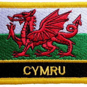 Könnte beinhalten: Ein roter Drache mit schwarzen Umrissen und gelben Akzenten auf einem weißen, grünen und schwarzen Hintergrund. Die Worte "CYMRU" sind in Gelb auf einem schwarzen Hintergrund geschrieben.