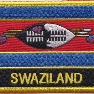 Può includere: Una toppa ricamata con la bandiera dello Swaziland. La toppa è rossa, gialla e blu con uno scudo nero e bianco e lance al centro. Il testo "SWAZILAND" è ricamato in giallo su sfondo nero.