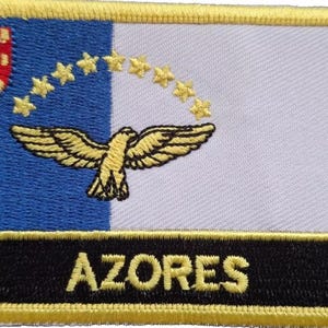 May include: A yellow embroidered patch with a blue and white flag depicting a yellow bird with outstretched wings and nine yellow stars. The text "AZORES" is embroidered in yellow on a black background.