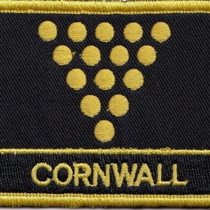 Könnte beinhalten: Ein gestickter schwarzer und goldener Aufnäher mit einer Gruppe goldener Kreise in Form einer Weintraube. Der Text "CORNWALL" ist in Gold unterhalb der Trauben gestickt.