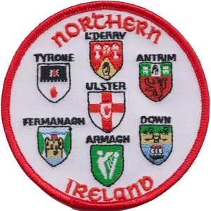 Puede incluir: Un parche circular blanco con un borde rojo. El texto "Northern Ireland" está escrito en rojo alrededor del borde. Dentro del círculo hay seis escudos con los escudos de armas de los seis condados de Irlanda del Norte: Antrim, Armagh, Down, Fermanagh, Londonderry y Tyrone. El escudo de Ulster está en el centro.