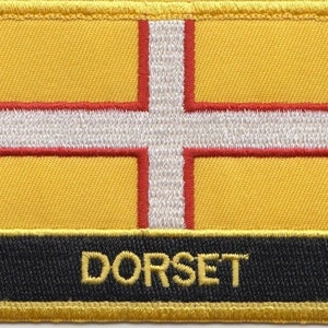 Op de afbeelding: Een gele geborduurde patch met een wit kruis en rode bies. De tekst "DORSET" is geborduurd in goud op een zwarte achtergrond.