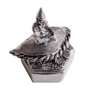 Puede incluir: Una figurita de estaño plateado de un gnomo sentado en un bote. El bote está sobre una base hexagonal con un diseño de cuerda alrededor del borde.