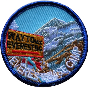 May include: A blue and white embroidered patch featuring a mountain scene with the text "WAY TO EVEREST BASE CAMP" in white and gold lettering. The text "WAY TO EVEREST" is on a yellow sign with a red border.